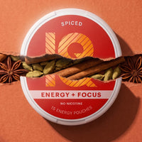 A round tin of Q10 Spiced Energy + Focus pouches, shown with a torn-paper effect revealing cinnamon sticks, cardamom pods, and star anise against a warm orange-brown background.